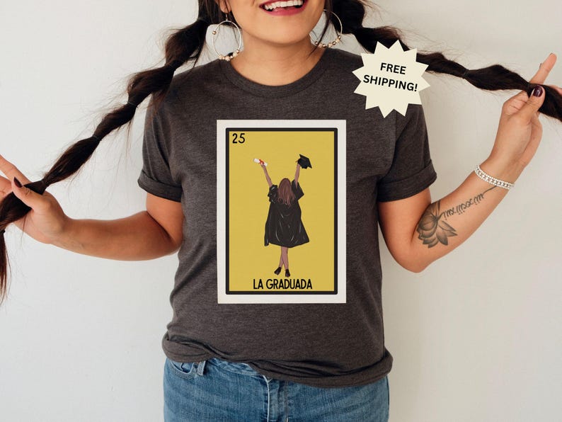 May include: A dark gray t-shirt with a graphic of a Loteria card featuring a woman in a graduation cap and gown. The card is labeled "25 La Graduada".