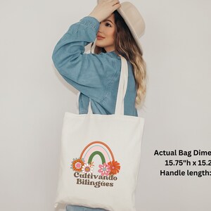Op de afbeelding: Een witte canvas tote bag met een regenboog- en bloemenontwerp. De tas heeft de tekst "Cultivando Biling&uuml;es" erop gedrukt. De tas is 40 cm hoog en 38,7 cm breed, met een handvatlengte van 54,6 cm.