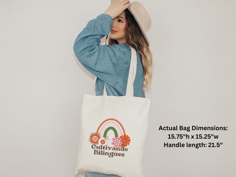 Op de afbeelding: Een witte canvas tote bag met een regenboog- en bloemenontwerp. De tas heeft de tekst "Cultivando Biling&uuml;es" erop gedrukt. De tas is 40 cm hoog en 38,7 cm breed, met een handvatlengte van 54,6 cm.