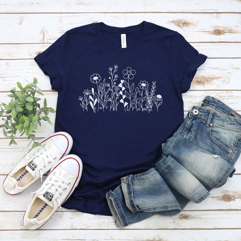 May include: A navy blue t-shirt with a white graphic of flowers and the text "Let It Bloom". The shirt is laid flat on a white wooden surface with a pair of white sneakers and blue jeans.