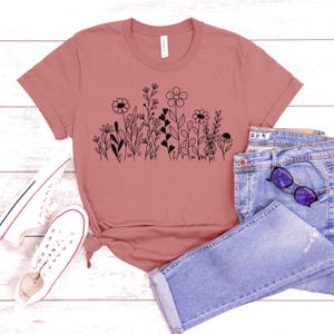 May include: A mauve t-shirt with a black floral design and the text "be kind" and "true north". The shirt is laying on a white surface with a pair of white sneakers and blue jeans.