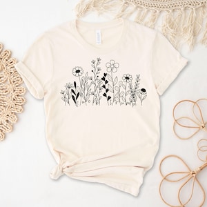 May include: A cream-colored t-shirt with a black floral design and the text "We are all just a little bit wild".