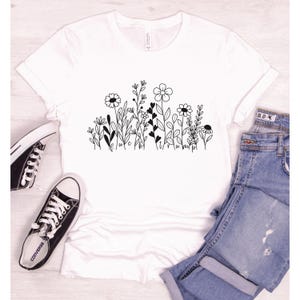 May include: White t-shirt with a black line drawing of a field of flowers. The flowers are stylized and have hearts in the stems. The text "luck" and "happy" are written in cursive at the bottom of the design.