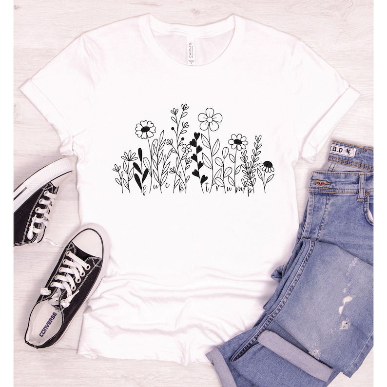 May include: White t-shirt with a black line drawing of a field of flowers. The flowers are stylized and have hearts in the stems. The text "luck" and "happy" are written in cursive at the bottom of the design.