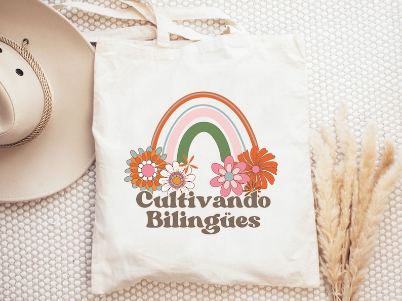 Op de afbeelding: Een witte tote bag met een regenboog en bloemen erop gedrukt. De tekst "Cultivando Biling&uuml;es" is onder de regenboog gedrukt.