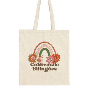 Op de afbeelding: Een witte tote bag met een regenboog- en bloemenprint. De tekst "Cultivando Biling&uuml;es" is onder de regenboog gedrukt.