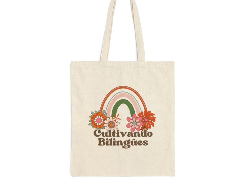 Op de afbeelding: Een witte tote bag met een regenboog- en bloemenprint. De tekst "Cultivando Biling&uuml;es" is onder de regenboog gedrukt.
