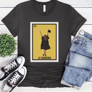 May include: A black t-shirt with a graphic of a Loteria card featuring a woman in a graduation cap and gown. The card is labeled "25 La Graduada".