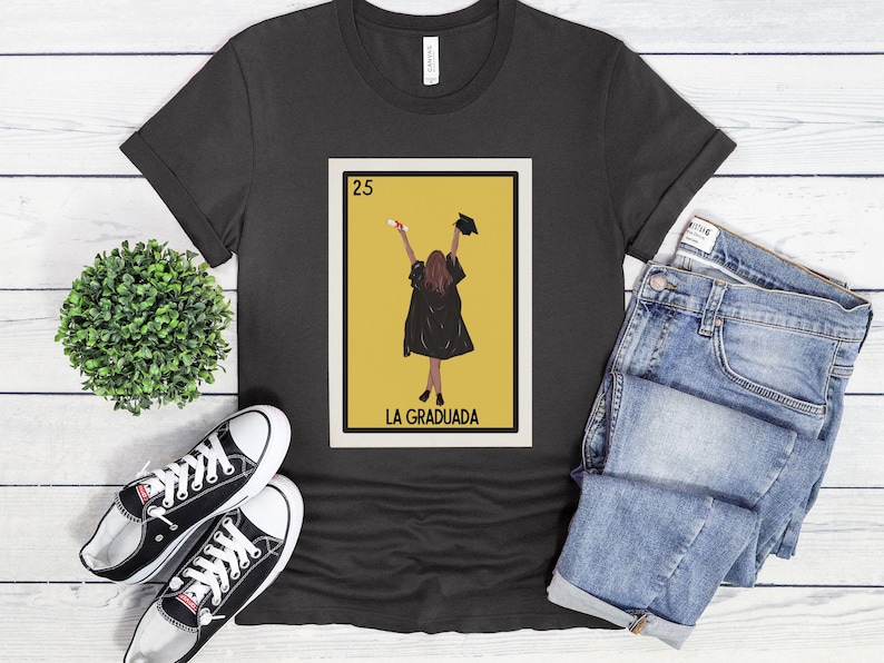 May include: A black t-shirt with a graphic of a Loteria card featuring a woman in a graduation cap and gown. The card is labeled "25 La Graduada".
