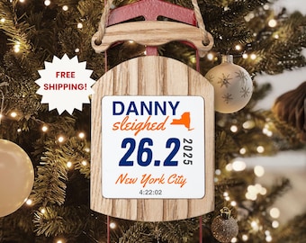 May include: Wooden sled-shaped Christmas ornament with a white rectangular insert. The insert reads "DANNY sleighed 26.2 2025 New York City 4:22:02" in blue and orange. The ornament hangs from a brown string and is set against a Christmas tree.