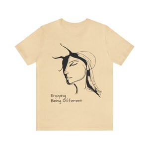 May include: A beige t-shirt with a black and white line drawing of a woman's face and the text "Enjoying Being Different".