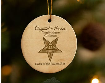 May include: A round, cream-colored Christmas ornament with a gold string for hanging. The ornament features the text "Cryuitel Mocker, Worthy Muster, Glesurwth" and "Order of the Eastern Star" with a star symbol. The ornament is from Rockn Pine Designs.