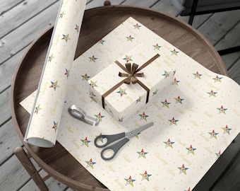 May include: Cream-colored wrapping paper with a repeating pattern of colorful stars, moons, and text. A wrapped gift box with a brown ribbon and bow sits on the paper. A roll of paper, scissors, and tape are also visible.
