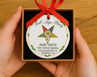 May include: A white, round Christmas ornament with a red ribbon, displayed in a black box. The ornament features a gold star with a smaller star inside, and the words "Faith, Hope, Charity" and "Mary Smith" are written on it. The ornament is decorated with holly.