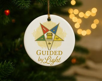 May include: A white ceramic Christmas ornament with a gold string. The ornament features a star design with various symbols and the text "GUIDED by Light" in gold lettering. The background is blurred with Christmas tree branches.