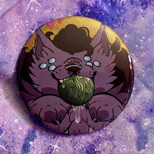 May include: A round, purple button featuring a cartoon wolf-like creature with multiple blue eyes, holding a green ball with a swirl design. The top of the button has gold glitter and pink hearts.