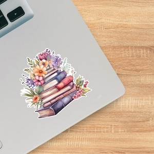 May include: A sticker featuring a stack of books in shades of blue, red, and cream, topped with a vibrant arrangement of purple, orange, and pink flowers with green leaves, displayed on the surface of a silver laptop.