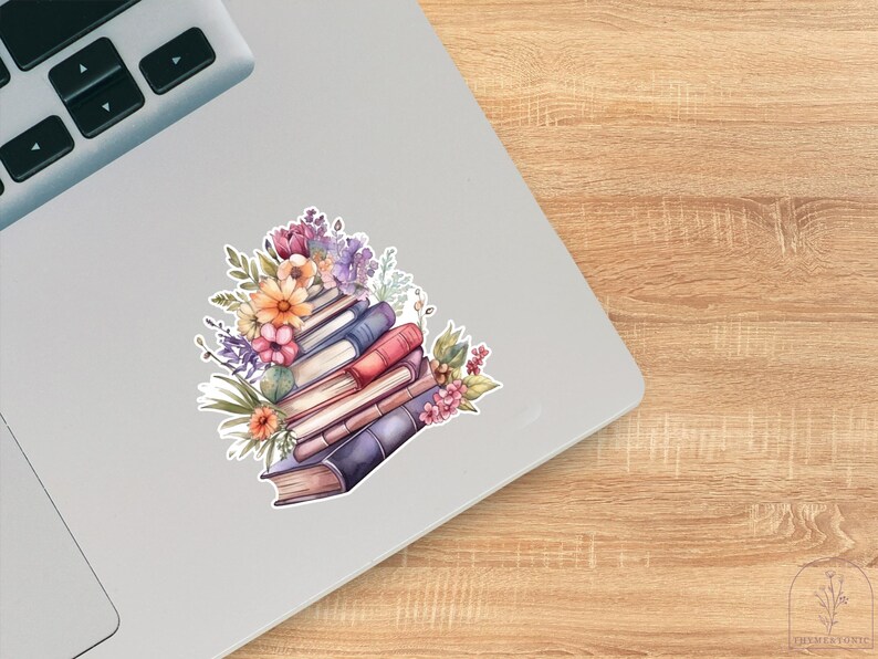 May include: A sticker featuring a stack of books in shades of blue, red, and cream, topped with a vibrant arrangement of purple, orange, and pink flowers with green leaves, displayed on the surface of a silver laptop.