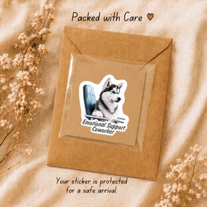 Pu&ograve; includere: Un adesivo raffigurante un husky bianco e nero seduto su una sedia da ufficio blu con il testo Emotional Support Coworker, in una busta di plastica trasparente su una busta di carta marrone con il testo Packed with Care e un'icona a cuore.