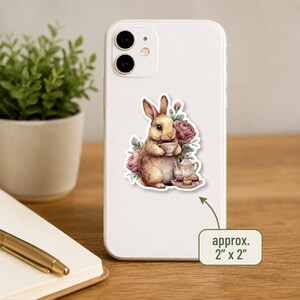 May include: A sticker on a white smartphone featuring a brown rabbit holding a teacup, surrounded by pink roses, a teapot, and small cakes. A green label with an arrow points to the sticker, stating the dimensions are approximately 2" x 2".
