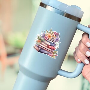 May include: A light blue insulated tumbler with a handle and a silver rim, featuring a sticker of a stack of books adorned with colorful flowers, including pink, orange, and purple blossoms, resting on the side of the beverage container.