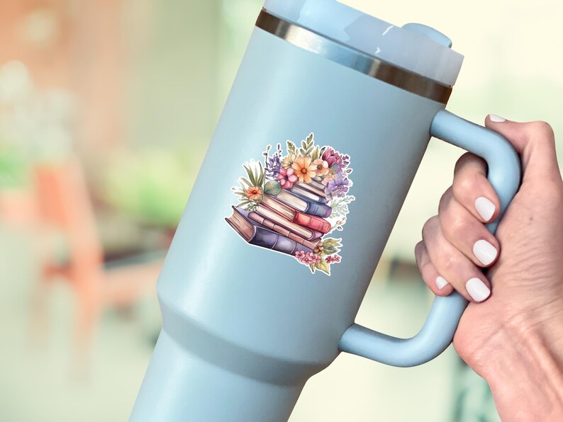 May include: A light blue insulated tumbler with a handle and a silver rim, featuring a sticker of a stack of books adorned with colorful flowers, including pink, orange, and purple blossoms, resting on the side of the beverage container.