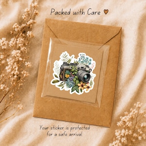 May include: A sticker featuring a vintage camera surrounded by colourful flowers and green leaves, enclosed in a clear protective sleeve on a brown paper envelope. The text Packed with Care and Your sticker is protected for a safe arrival.