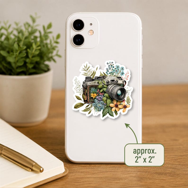May include: A white smartphone featuring a sticker of a vintage camera surrounded by colourful flowers and green foliage. The sticker measures approximately 2 inches by 2 inches, as indicated by the text on the image.