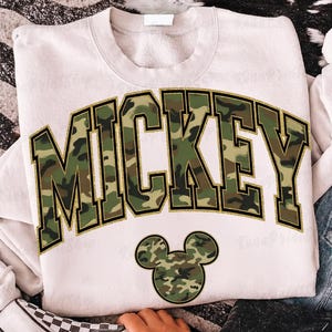 以下が含まれることがあります： 「MICKEY」の文字が大きく迷彩柄で金色の縁取りが施された白いスウェットシャツ。テキストの下には、お揃いのミッキーマウスの頭のグラフィックがあります。スウェットシャツはクルーネックです。