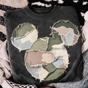 May include: Dark gray sweatshirt featuring a patchwork design of a mouse head. The mouse head is made of various fabric patches in shades of green, pink, blue, and cream. The sweatshirt has a crew neck and long sleeves.