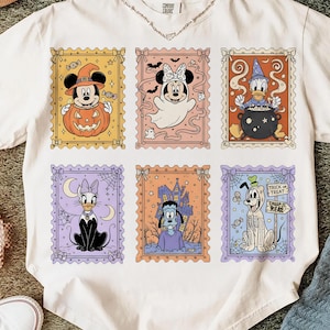 May include: White t-shirt featuring six Halloween-themed postage stamp designs with Disney characters. The stamps showcase Mickey Mouse, Minnie Mouse, Donald Duck, Daisy Duck, Goofy, and Pluto in spooky costumes. The shirt has a crew neck and short sleeves.