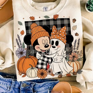 May include: Cream-colored sweatshirt featuring Mickey Mouse and a ghost embracing, both wearing orange hats. The design includes pumpkins, flowers, and fall leaves. The background has a black and white plaid pattern. A tan hat and blue jeans are visible.