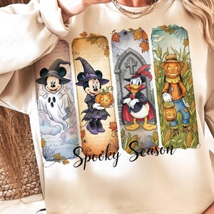 May include: Beige sweatshirt featuring four Halloween-themed Disney character illustrations. Mickey Mouse as a ghost, Minnie Mouse with a pumpkin, Donald Duck as a vampire, and a scarecrow. The words "Spooky Season" are written below.