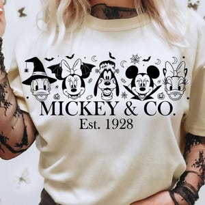 May include: Cream-colored t-shirt featuring a black and white graphic of Disney characters in Halloween costumes. The text "MICKEY & CO. Est. 1928" is printed below the characters. The shirt has short sleeves.