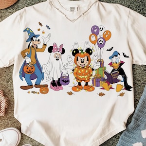 May include: White t-shirt featuring a Halloween-themed graphic with Disney characters. Goofy, Minnie Mouse, and Mickey Mouse are dressed in costumes. The design includes pumpkins, ghosts, and balloons, with the text "Spooky Tales" on a book.