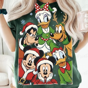 May include: Green t-shirt featuring a festive graphic of Disney characters Mickey Mouse, Minnie Mouse, Goofy, Donald Duck, and Daisy Duck in Santa hats and Christmas attire. The design includes snowflakes, creating a holiday theme.