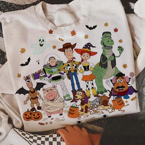 May include: Off-white sweatshirt featuring a Halloween-themed design with cartoon characters. The design includes a ghost, bats, pumpkins, and various Toy Story characters dressed in Halloween costumes. The sweatshirt has a crew neck and long sleeves.