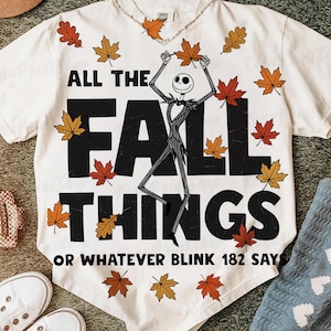 May include: White t-shirt with the text "ALL THE FALL THINGS" and a Jack Skellington character illustration. The shirt is decorated with autumn leaves in shades of red, orange, and yellow. The phrase "OR WHATEVER BLINK 182 SAY" is printed at the bottom.