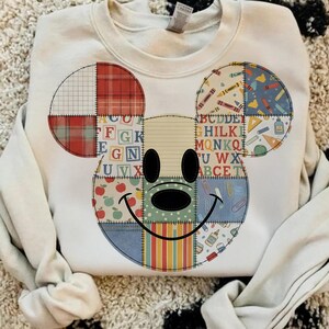 Könnte beinhalten: Cremefarbenes Sweatshirt mit einem Patchwork-Design eines lächelnden Gesichts. Das Design enthält Karomuster, Buchstaben und Schulmaterialien. Das Gesicht hat schwarze ovale Augen und eine schwarze Nase. Das Sweatshirt hat lange Ärmel.