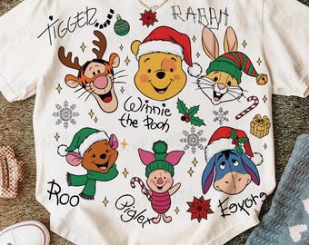 May include: White t-shirt featuring cartoon characters from Winnie the Pooh, including Tigger, Winnie, Rabbit, Roo, Piglet, and Eeyore, all wearing Christmas hats. The shirt has the words "Tigger", "Rabbit", "Winnie the Pooh", "Roo", "Piglet", and "Eeyore" printed on it.