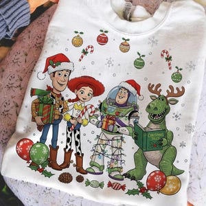 May include: White sweatshirt featuring Toy Story characters Woody, Jessie, Buzz Lightyear, and Rex, all wearing Santa hats. The design includes Christmas ornaments, candy canes, and the text "Christmas Tales" on a book.