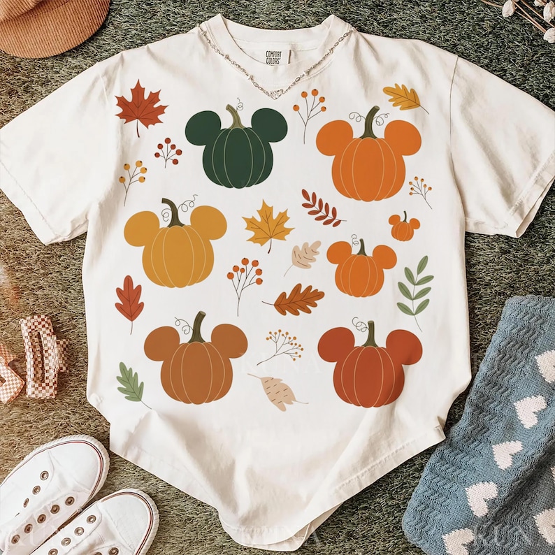 May include: Cream-colored t-shirt featuring a whimsical fall-themed design. The shirt showcases various pumpkins in shades of orange, brown, and green, along with autumn leaves and floral accents. The shirt has a relaxed fit and short sleeves.