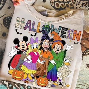 K&ouml;nnte beinhalten: Ein cremefarbenes Halloween-Sweatshirt mit Comicfiguren in Kost&uuml;men. Das Wort "HALLOWEEN" ist in bunten, dekorativen Buchstaben aufgedruckt. K&uuml;rbisse, Flederm&auml;use und Kerzen sind ebenfalls im Design enthalten.
