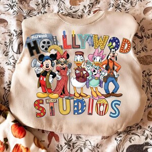 K&ouml;nnte beinhalten: Beiges Sweatshirt mit einer farbenfrohen Grafik von Disney-Figuren, darunter Micky und Minnie Maus, Donald Duck, Daisy Duck und Goofy. Der Text "HOLLYWOOD STUDIOS" wird in einer lebendigen, cartoonartigen Schriftart mit einer Filmrollen-Grafik angezeigt.