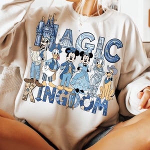 K&ouml;nnte beinhalten: Cremefarbenes Sweatshirt mit den Worten "MAGIC KINGDOM" in blauen und beigen Buchstaben. Das Design zeigt Illustrationen von Disney-Figuren wie Mickey Mouse, Minnie Mouse, Donald Duck und Goofy, alle in blauen Outfits.