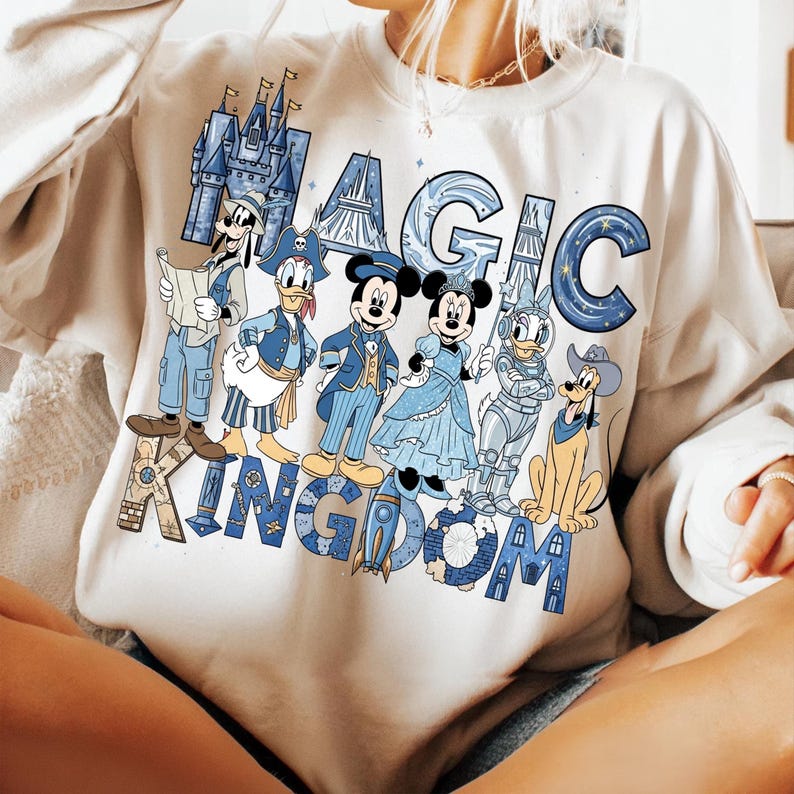 K&ouml;nnte beinhalten: Cremefarbenes Sweatshirt mit den Worten "MAGIC KINGDOM" in blauen und beigen Buchstaben. Das Design zeigt Illustrationen von Disney-Figuren wie Mickey Mouse, Minnie Mouse, Donald Duck und Goofy, alle in blauen Outfits.