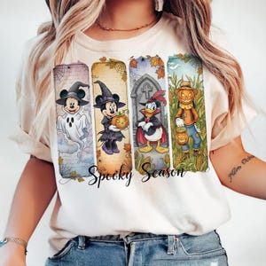 May include: Cream-colored t-shirt featuring a Halloween-themed design. The design includes four vertical panels with Disney characters dressed in costumes, including Mickey Mouse as a ghost and Minnie Mouse as a witch. The words "Spooky Season" are written in a script font.