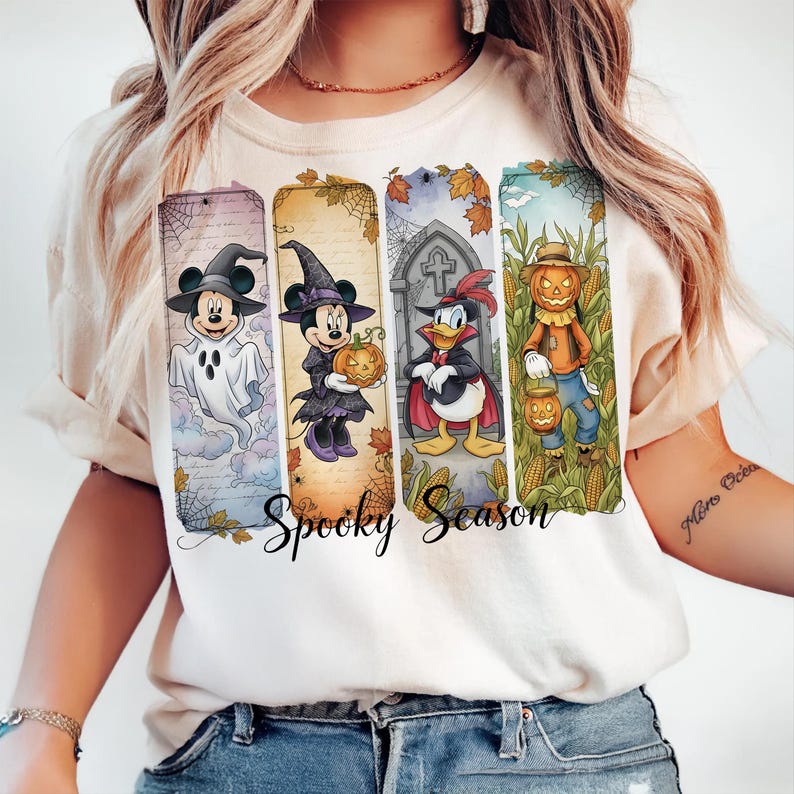 May include: Cream-colored t-shirt featuring a Halloween-themed design. The design includes four vertical panels with Disney characters dressed in costumes, including Mickey Mouse as a ghost and Minnie Mouse as a witch. The words "Spooky Season" are written in a script font.