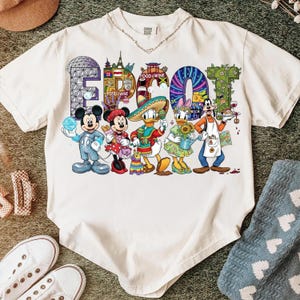 K&ouml;nnte beinhalten: Cremefarbenes T-Shirt mit dem Wort "EPCOT" in bunten Buchstaben und Disney-Figuren. Micky, Minnie, Donald, Daisy und Goofy sind abgebildet. Das Shirt hat einen Rundhalsausschnitt und kurze &Auml;rmel.