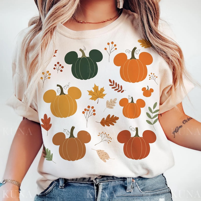 May include: Beige t-shirt featuring a fall-themed design. The design includes pumpkins with mouse ears in various colors, such as orange, green, and brown, along with autumn leaves and sprigs. The shirt is a casual, everyday wear item.